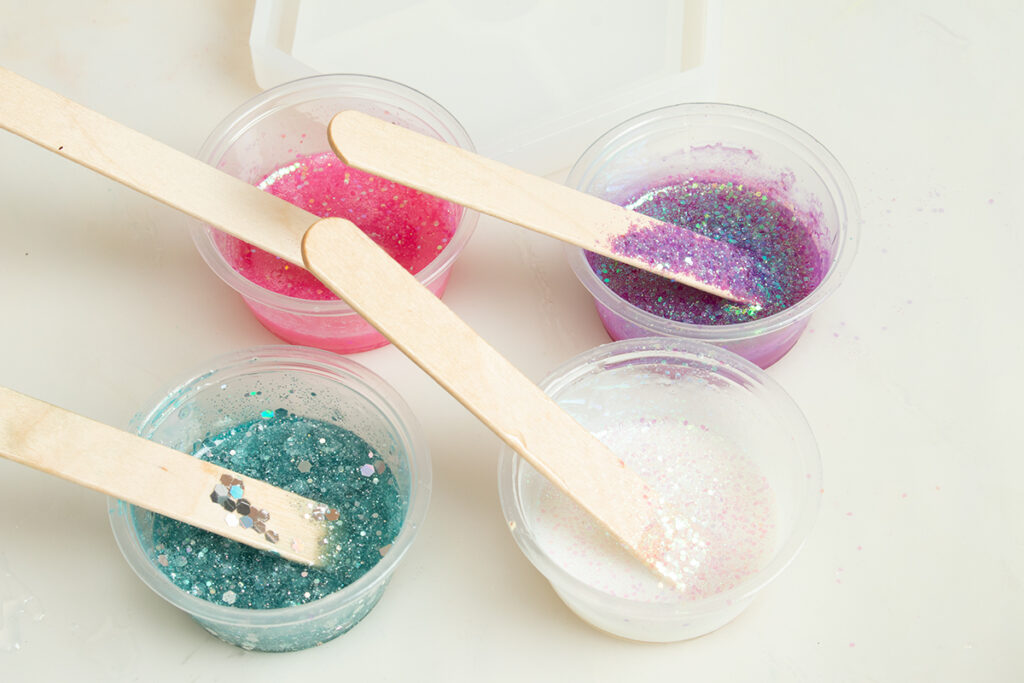 Four small cups of glittery resin with popsicle sticks sticking out of them. The resin colors are pink, purple, blue, and white.