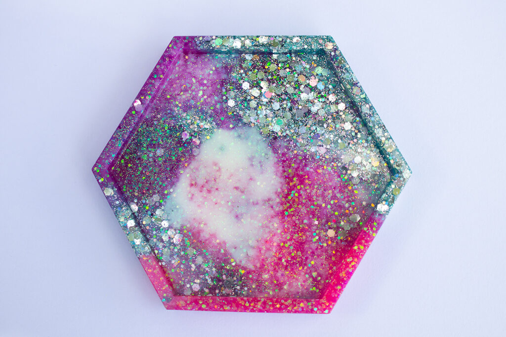 Fully cured epoxy resin hexagon-shaped coaster. The coaster is pink, purple, blue, and white with iridescent and silver holographic glitter.