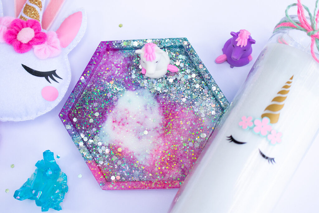 White felt unicorn face ornament with pink flowers, glitter epoxy resin coaster that is pink, purple, blue, and white, tall white candle with unicorn face on a white background