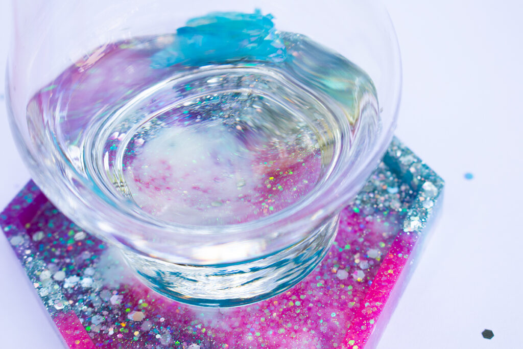 Clear glass sitting on top of a fully cured epoxy resin hexagon-shaped coaster. The coaster is pink, purple, blue, and white with iridescent and silver holographic glitter.